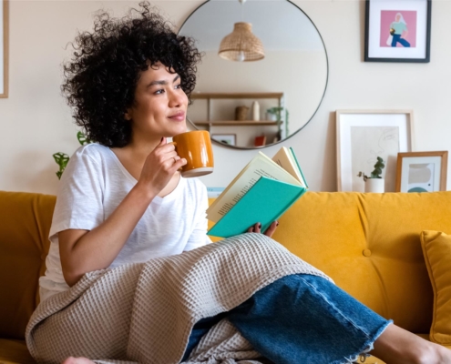 Image of a person sitting on the couch while reading a book and drinking coffee.